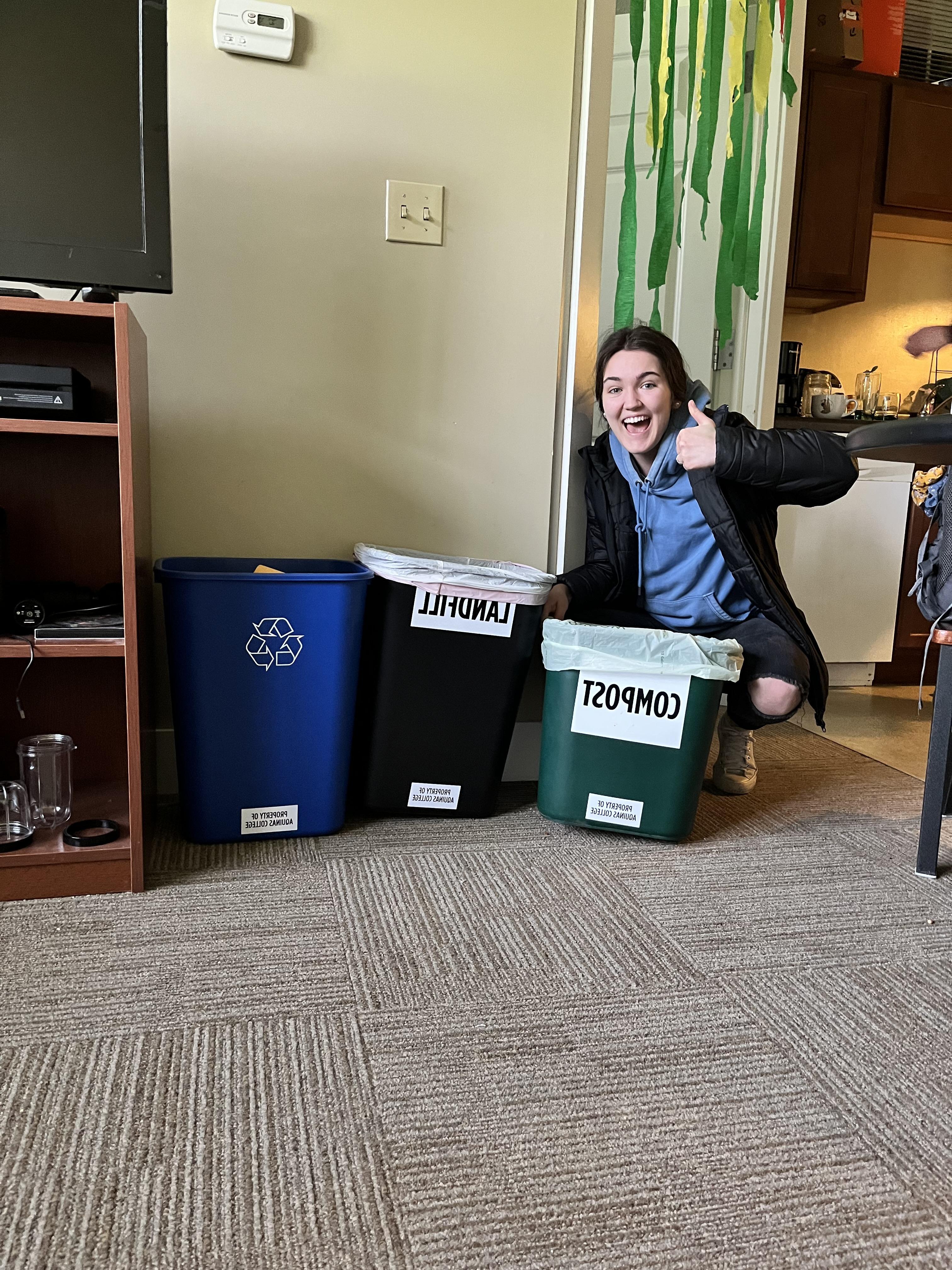 Student giving a thumbs up and sitting on the floor of their home or dorm with three bins labeled compost, landfill, and recycling. 