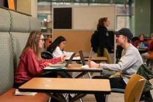 Two students sitting across from one another at a table smiling with their laptops open