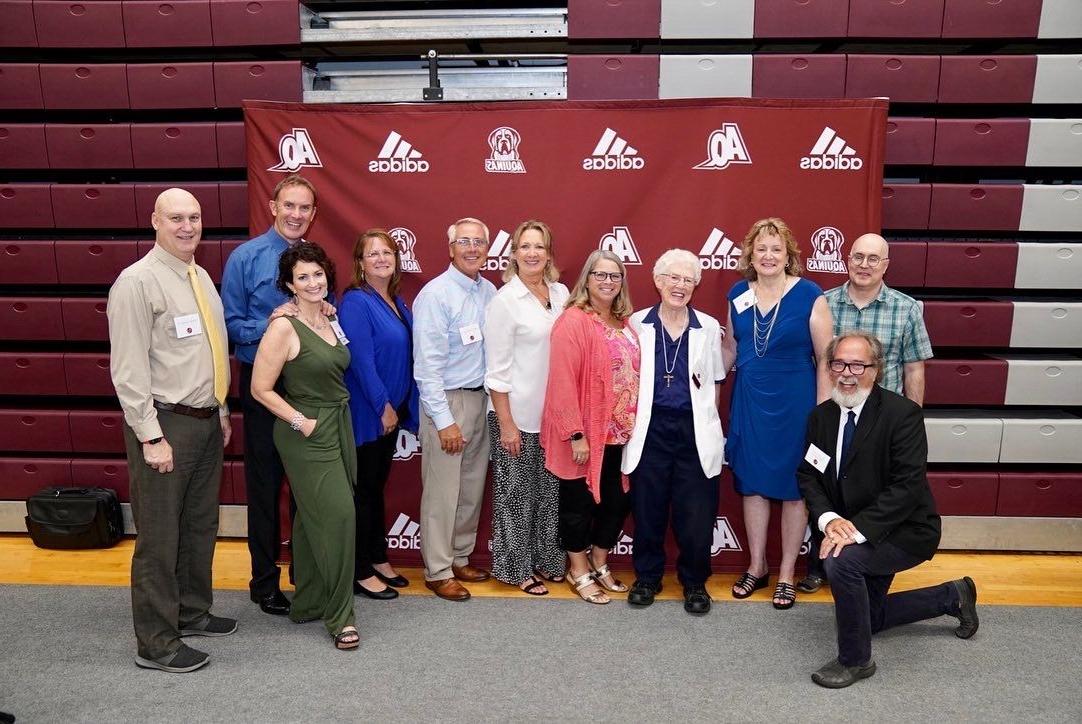 A group of smiling people, including Sister Ann Mason, posing for a photo in front of a Red AQ/Adidas printed backdro
