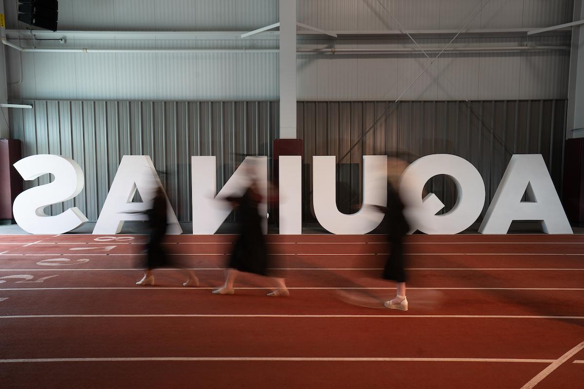 Large Aquinas letters set up on a track with blurred motion students in graduation caps and gowns walking in front