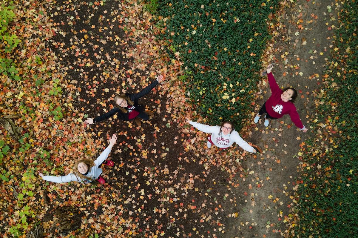 Overhead shot of three students with arms outstretched on a leafy trail in fall smiling up at the camera